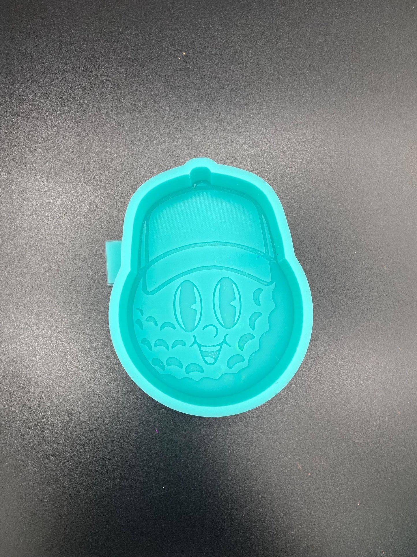 May include: Teal silicone mold of a cartoon golf ball wearing a baseball cap. The golf ball has a smiling face with simple eyes and a wide mouth. The mold is designed for creating resin crafts or other similar projects. The mold is a bright teal color and shows fine details of the golf ball texture and facial features. The baseball cap sits atop the golf ball, adding to the whimsical design.
